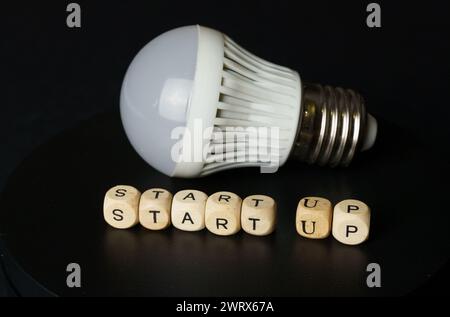 Eine Glühbirne liegt neben hölzernen Buchstabenblöcken, die das START-UP beschreiben und den Beginn eines neuen Unternehmens symbolisieren. Stockfoto