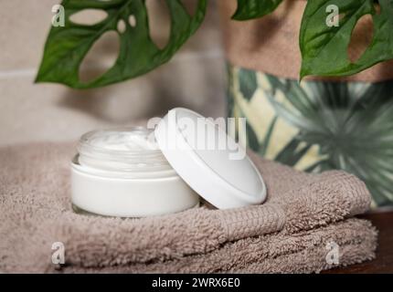 Geöffnetes Cremeglas mit leerem Deckel auf braunem gefaltetem Handtuch in der Nähe von grüner Monstera-Pflanze auf hölzernem Badezimmer-Arbeitsplatte, Nahaufnahme, Kosmetikverpackungsmotiv Stockfoto