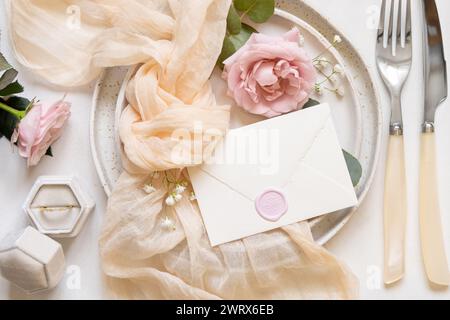 Versiegelter Umschlag in der Nähe von Knoten aus cremefarbenem Tüll und hellrosa Rosen auf den Tellern von oben, Kopierraum. Hochzeit Schreibwaren Mockup mit Verlobungsring in A Stockfoto