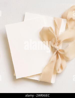 Karten, die mit einem beigefarbenen Seidenband gebunden sind, auf der weißen Tischplatte. Hochzeit Schreibwaren Mockup. Romantische Szene mit vertikalen leeren Karten. Hochzeit, Valentinstag, Stockfoto