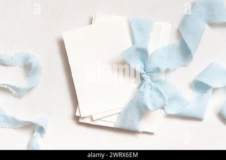 Karten, die mit hellblauen Seidenbändern gebunden sind, auf weißem Tisch. Hochzeit Schreibwaren Mockup. Romantische Szene mit vertikalen leeren Karten. Hochzeit, Valen Stockfoto