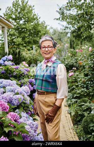 Debonair fröhliche reife Frau mit Brille, die sich in der Nähe von leuchtenden Hortensien posiert und in die Kamera blickt Stockfoto