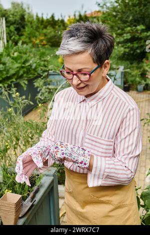 Fröhliche, schöne reife Frau mit Brille und Handschuhen, die in ihrem Garten posiert und wegblickt Stockfoto