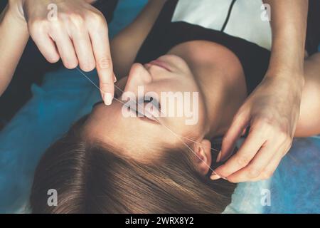 Die Make-up-Künstlerin zupft ihre Augenbrauen mit einem Faden einer schönen Brünette in einem Schönheitssalon. Professionelle Gesichtspflege. Stockfoto