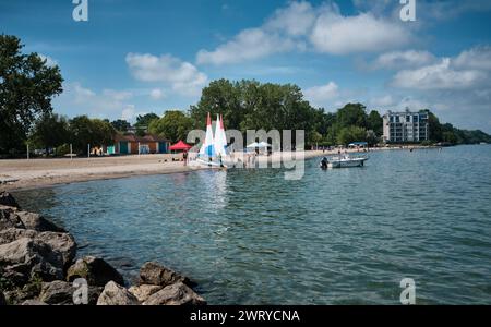 Farbenfrohe rot-weiß-blaue Segel auf Segelbooten am Sandstrand mit Schwimmern und Sonnenanbetern am Lakeside Park Beach, Port Dalhousie, Ontario. Stockfoto