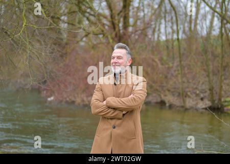 Mann mittleren Alters mit grauen Haaren und grauem Bart in einem Park im Winter oder Frühling mit gekreuzten Armen Stockfoto