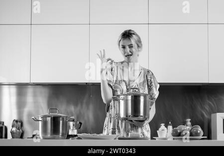 Eine Frau mit Kochtopf steht neben dem Tisch in der Küche zu Hause. Die Hausfrau kocht morgens in der Küche. Stockfoto