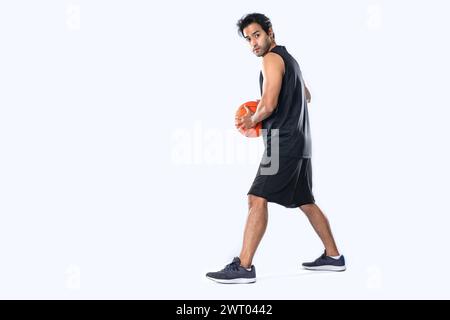 Männlicher Basketballspieler in Sportbekleidung, der Basketball auf weißem Hintergrund mit einem bestimmten Gesicht hält. Stockfoto