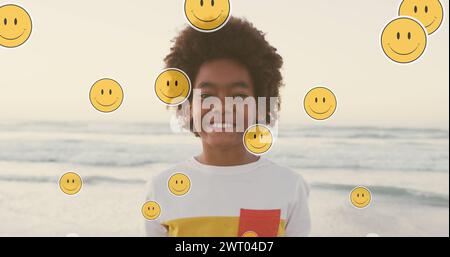 Bild von Emoji-Ikonen über einem glücklichen afroamerikanischen Jungen am sonnigen Strand Stockfoto