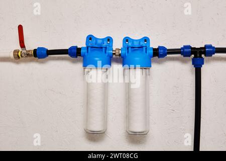 Das Wasserfiltersystem mit zwei durchsichtigen Kanistern und blauen Deckeln ist an der weißen Wand montiert, die durch schwarze und blaue Leitungen verbunden ist, wobei das rote Absperrventil eingeschaltet ist Stockfoto