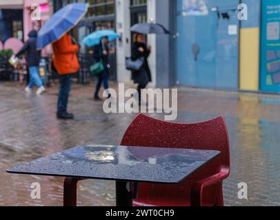 Londoner Fußgängerzone mit regengetränktem Tisch und Stuhl im Vordergrund. Leute mit Regenschirmen laufen im Regen im Hintergrund. Stockfoto