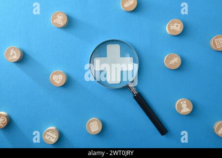 Medizin- und Versicherungskonzept mit Vergrößerungsglas, Kreuz und Holzmarken, die verschiedene medizinische Leistungen auf blau isoliertem bac zeigen Stockfoto