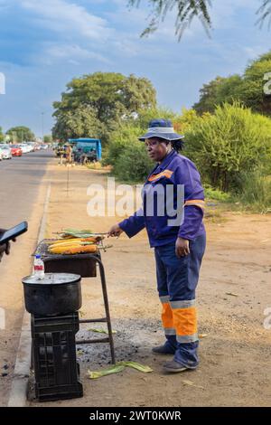 afrikanische Frau Straßenverkäuferin, die gegrilltes Getreide auf einer belebten Straße in der Stadt auf dem Bürgersteig verkauft, Autos im Hintergrund Stockfoto