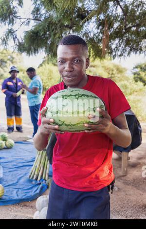 afrikanischer Straßenverkäufer, der Wassermelonen am Straßenrand verkauft, Kunden im Hintergrund Stockfoto