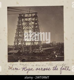 Drahtseil über den Fall. Unbekannt, Fotograf 1868–1869 View of the Falls View Suspension Bridge, auch bekannt als Niagara Clifton Bridge, im Bau. Eine kleine Hütte ist vor einem der Pylontürme zu sehen. Das Deck der Brücke erstreckt sich in die Entfernung. (Recto, Mount) unten links, unten Bild, in Bleistift: „Drahtseil quer durch den Fall“; unten links, unten Bild, in violetter Tinte: „Drahtseil quer durch den Fall“; Stockfoto