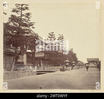 Eingangstor eines nicht identifizierten japanischen Tempels. Unbekannt, Fotograf aus den 1850er–1890er Jahren Blick auf den Vordereingang zu einem nicht identifizierten japanischen Tempel oder Schrein. Eine Brücke führt zum Eingangstor und überquert einen schmalen Graben. Standlaternen säumen den Graben auf der anderen Seite der Brücke. Zwei Männer stehen auf dem Feldweg, der das Eingangstor passiert, vor der Kamera, während ein dritter unter dem Tor steht, mit einem Ventilator, der sein Gesicht vor der Sonne schützt. Ein zweites, kleineres Gate steht im Hintergrund auf der rechten Seite des Bildes. Stockfoto