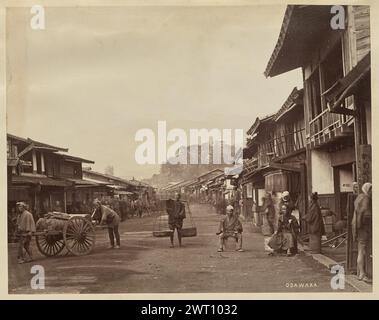 Odawara. Baron Raimund von Stillfried, Fotograf (Österreich, 1839 - 1911) 1877–1880 Blick auf eine Straße in Odawara, in der japanischen Präfektur Kanagawa. Geschäfte säumen beide Seiten der unbefestigten Straße, die von Fußgängern und Händlern wimmelt. Ein Mann steht mitten auf der Straße, der Kamera zugewandt, mit zwei Körben, die an einem Tragemast befestigt sind, über seinen Schultern, während zwei Männer an ihm vorbeigehen und einen Holzwagen schieben, der mit Holzbündeln und Holzzündern beladen ist. Unten rechts, handgeschrieben im negativ, in weiß: 'ODAWARA.'; handgeschrieben im negativ, in Blac Stockfoto