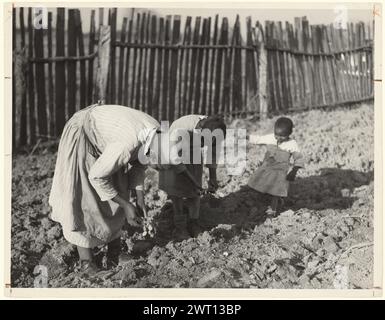 Bei der Arbeit in Garden, Gee's Bend, Alabama. Arthur Rothstein, Fotograf (Amerikaner, 1915–1985) 1937 Eine Frau und ein Mädchen bücken sich, während sie im Garten Setzlinge Pflanzen. Ein junges Mädchen geht hinter ihnen vorbei. Der Garten ist von einem Holzzaun umgeben. Stockfoto