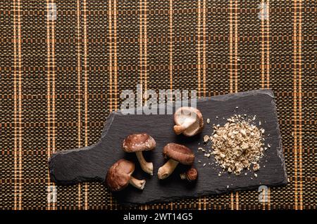 Flache Ansicht von trockenem Pulver aus Shiitake-Pilzen, Lentinula edodes. Lebensmittelzutat auf Schneidebrett aus schwarzem Stein mit frischen Shiitake-Pilzen. Stockfoto