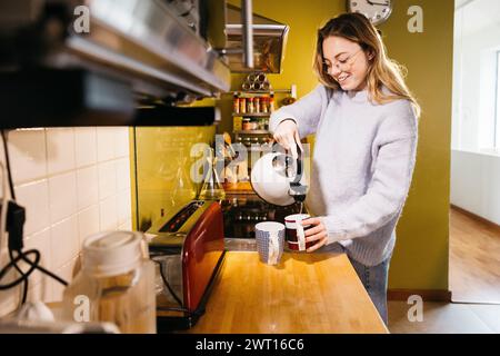 Eine junge, schöne Frau, die morgens heißes Wasser in einen Becher in der Küche gießt. Junge Frau, die Tee zum Frühstück in einer Küche zubereitete. Stockfoto