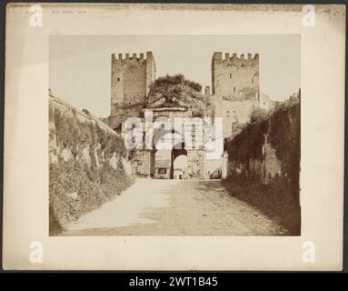 Bogen von Drusus. Unbekannt, Fotograf um 1865–1880 ein bogenförmiges Steintor, flankiert von zwei gleich hohen Türmen. Die Oberseite jedes Turms ist mit Zinnen versehen. Gras wächst oben auf dem Bogen. Die Wände entlang der Straße, die zum Bogen führt, sind mit Gras und Efeu bedeckt. (Recto, Mount) oben links, blau getippter Druck auf Papieretikett: "B.I. 78 Old Roman Gate"; (Verso, Mount) oben links, schwarze Tinte: "Old Roman Gate"; Stockfoto