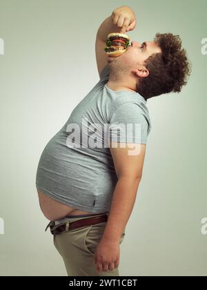 Fast Food, hungrig und Mann mit Hamburger im Studio Hintergrund mit Mittagessen, Snack und Verlangen nach einem Mann mit einer großen Größe. Männlicher Mensch, Burger und zum Mitnehmen Stockfoto