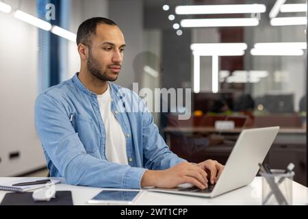 Konzentrierter und seriöser junger hispanischer Mann, der in einem modernen Büro arbeitet, an einem Schreibtisch sitzt und auf einem Laptop schreibt. Stockfoto