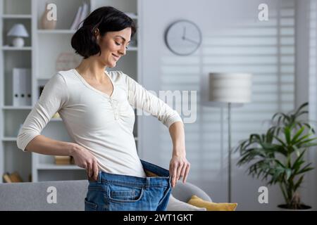 Eine fröhliche Frau zu Hause zeigt ihre erfolgreiche Gewichtsreduktion, indem sie eine alte, übergroße Jeans anprobiert und zufrieden lächelt. Stockfoto