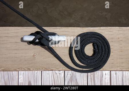 Ein schwarzes Festmacherseil mit einem geknüpften Ende, das um eine Klampe auf einem hölzernen Pier gebunden ist. Stockfoto