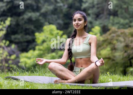 Lächelnde und entspannte junge Indianerin in Sportbekleidung, die im Park auf einer Matte in Lotusposition sitzt und Yoga macht, meditiert und erholt. Stockfoto