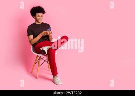 Ganzkörperfoto eines entspannten jungen Mannes, der mit dem Smartphone sitzt, überprüft seine E-Mail-Nachrichten isoliert auf rosa Hintergrund Stockfoto