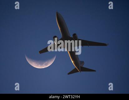 Ein kommerzielles Flugzeug in niedriger Höhe kommt bei Sonnenuntergang an einem Halbmond vorbei. Stockfoto