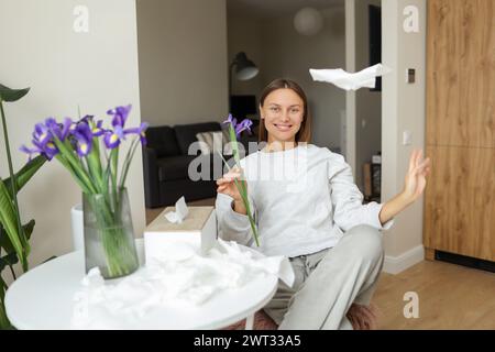 Allergiefrei. Eine fröhliche junge Frau wirft zu Hause Servietten hoch. Glückliches Leben ohne laufende Nase, Juckreiz oder Husten Symptome. Allergenstrauß von Irisblumen in der Vase, benutzte Taschentücher auf dem Tisch. Stockfoto