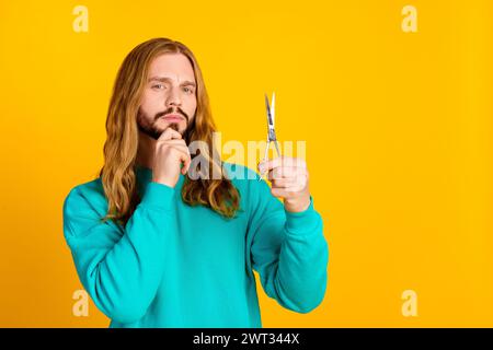 Porträt der gesinnten nachdenklichen Person Handberührung Kinn halten Schere betrachten leeren Raum isoliert auf gelbem Hintergrund Stockfoto