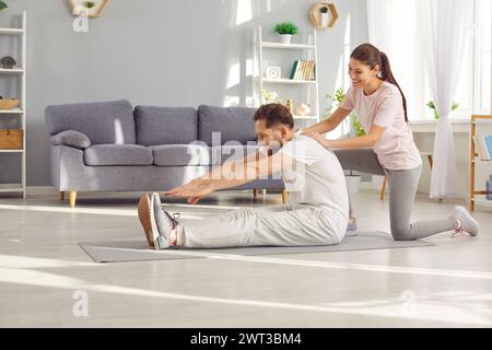 Glückliche, sportliche junge Frau, die ihrem Mann bei Sportübungen zu Hause hilft Stockfoto