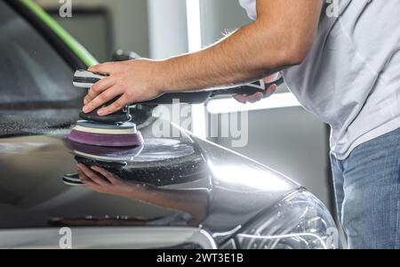 Junger Mann, der die Motorhaube des Autos mit einem Orbitalpolierer poliert, um Kratzer am Auto zu entfernen, detailliert das Konzept Stockfoto