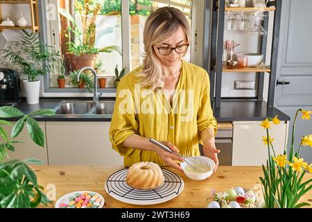 Blonde Frau, die Glasur für Osterkuchen in der Küche vorbereitet. Traditioneller osterkuchen oder süßes Brot mit Belag. Ostervergnügen. Stockfoto