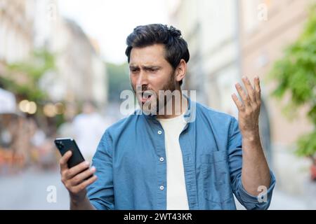 Ein wütender junger Mann in legeren Kleidern steht auf einer Straße der Stadt und schaut besorgt auf den Bildschirm des Telefons und winkt inkohärent mit den Händen. Stockfoto