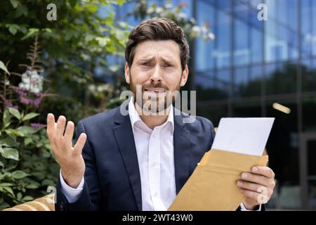 Nahporträt eines traurigen und enttäuschten jungen Geschäftsmannes, der draußen sitzt, einen offenen Umschlag mit einem Brief in der Hand hält und in die Kamera blickt. Stockfoto