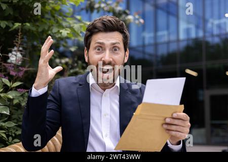 Nahaufnahme eines glücklichen und schockierten jungen Mannes in einem Business-Anzug, der draußen in einem Bürogebäude sitzt, einen gerissenen Umschlag mit einem Brief in der Hand hält und vor Freude die Kamera anschrie. Stockfoto