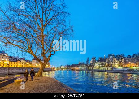 Ein Paar genießt einen Spaziergang in der Dämmerung entlang der seine, während das Ambiente von Paris um sie herum leuchtet. Stockfoto