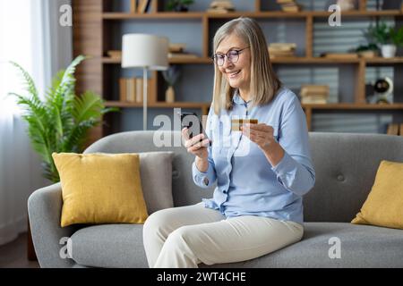 Fröhliche reife Frau, die auf dem Sofa sitzt, ihr Smartphone benutzt und eine Kreditkarte hält, lächelt, während sie in einem gemütlichen Wohnzimmer online shoppt. Stockfoto