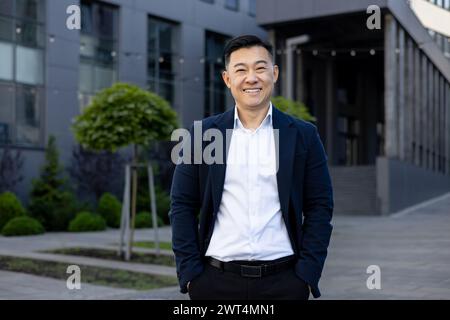 Porträt eines lächelnden asiatischen Geschäftsmannes in einem Anzug, der an einem sonnigen Tag selbstbewusst vor einem modernen Bürokomplex steht. Stockfoto