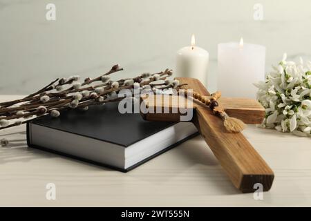 Brennende Kirchenkerzen, Holzkreuz, Rosenkranzperlen, Bibel, Weidenzweige und Schneeglöckchen auf weißem Tisch Stockfoto