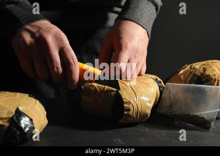 Schmuggel und Drogenhandel. Mann, der ein Paket mit Betäubungsmitteln öffnet, mit einem Cutter am dunklen Tisch, Nahaufnahme Stockfoto