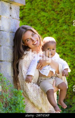 Glückliche junge Mutter, vielfältige Eltern mit Baby im Sommerhof. Überfröhliche Mutter mit einem kleinen Kind, das einen ethnischen oder multirassischen Hintergrund hat Stockfoto