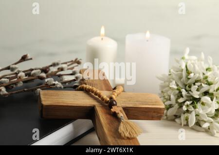 Brennende Kirchenkerzen, Holzkreuz, Rosenkranzperlen, Bibel, Weidenzweige und Schneeglöckchen auf weißem Tisch, Nahaufnahme Stockfoto