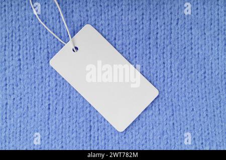 Leeres Bekleidungsetikett auf blauem Strickpullover-Hintergrund. Modell, Vorlage mit leerer rechteckiger Karte, Stoffanhänger für Preis oder Text. Shopping, Sale, Di Stockfoto