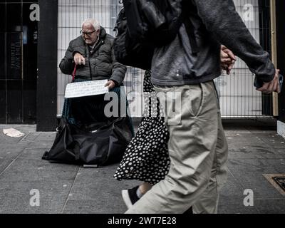 Ein Paar, das Hände hält, geht an einem behinderten alten Mann (möglicherweise obdachlos) vorbei, der auf den Straßen von London bettelt und ein Plakat hält, das um Hilfe bittet. Stockfoto