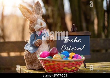 16. März 2024: Frohe Ostergrüße auf einem Schild neben einem Osterhasen mit Osternest und bunten Eiern. FOTOMONTAGE *** Frohe Ostern Grüße zum Osterfest auf einem Schild neben einem Osterhasen mit Osternest und bunten Eiern. FOTOMONTAGE Stockfoto